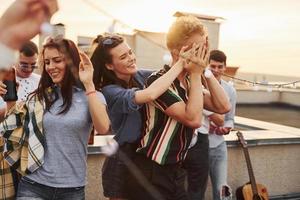 Playing game. Man's eyes covered by hands. Group of young people in casual clothes have a party at rooftop together at daytime photo