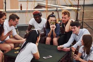 Sitting by table and playing card game. Group of young people in casual clothes have a party at rooftop together at daytime photo