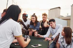 Sitting by table and playing card game. Group of young people in casual clothes have a party at rooftop together at daytime photo