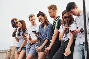 de pie con teléfonos y alcohol en las manos. un grupo de jóvenes con ropa informal tienen una fiesta en la azotea juntos durante el día foto