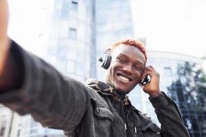 Listening to music in headphones. Young african american man in black jacket outdoors in the city standing against modern business building photo