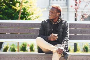 With phone in hand. Young african american man in black jacket outdoors in the city sitting on bench photo