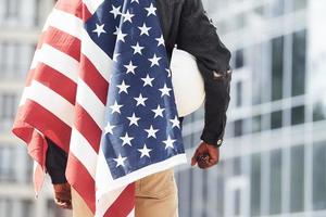 View from behind. Patriot holding USA flag. Conception of pride and freedom. Young african american man in black jacket outdoors in the city standing against modern business building photo