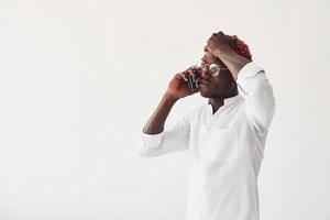Young african american man in white formal clothes talking by the phone against wall indoors photo