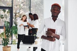 Man with notepad standing in front of colleagues. Group of african american business people working in office together photo