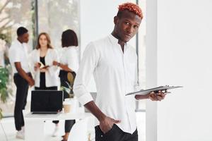 Man with notepad standing in front of colleagues. Group of african american business people working in office together photo