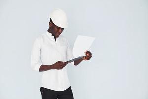 Young african american engineer in white formal clothes standing against wall indoors with documents photo