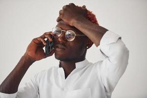 Young african american man in white formal clothes talking by the phone against wall indoors photo