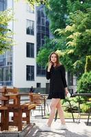 Talking by phone and holding cup of drink. Happy young girl in black skirt outdoors in the city near green trees and against business building photo