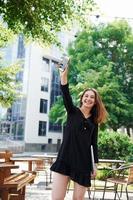 Holding laptop and phone. Happy young girl in black skirt outdoors in the city near green trees and against business building photo