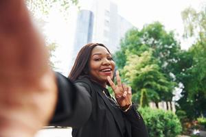 Doing selfie. Young afro american woman in fashionable clothes outdoors in the city near green trees and against business building photo