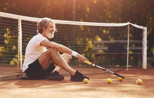 Sitting and taking a break. Senior stylish man in white shirt and black sportive shorts on tennis court photo