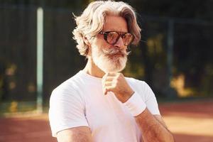 hombre mayor con estilo en anteojos, camisa blanca y pantalones cortos deportivos negros en la cancha de tenis foto