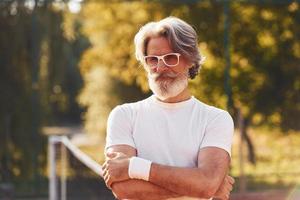 hombre mayor con estilo en anteojos, camisa blanca y pantalones cortos deportivos negros en la cancha de tenis foto