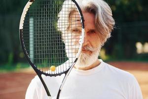 sosteniendo la raqueta. Senior hombre moderno y elegante con raqueta al aire libre en la cancha de tenis durante el día foto