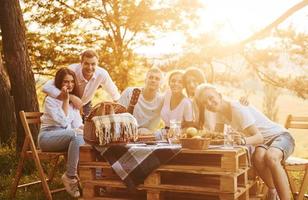 Sitting by picnic table. Group of young people have vacation outdoors in the forest. Conception of weekend and friendship photo