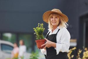Holding pot in hands. Senior woman is in the mini garden at daytime. Building exterior behind photo