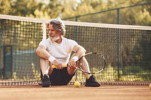 sentado y tomando un descanso. hombre mayor con estilo en camisa blanca y pantalones cortos deportivos negros en la cancha de tenis foto