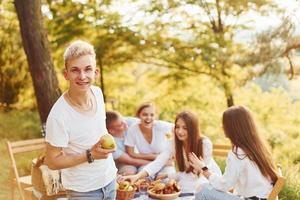 Positive guy with apple standing in front of people. Group of young people have vacation outdoors in the forest. Conception of weekend and friendship photo