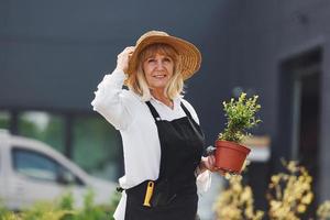 Holding pot in hands. Senior woman is in the mini garden at daytime. Building exterior behind photo