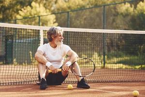 Sitting and taking a break. Senior stylish man in white shirt and black sportive shorts on tennis court photo