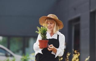 Holding pot in hands. Senior woman is in the mini garden at daytime. Building exterior behind photo