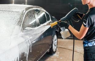 utilizando agua a alta presión. el automóvil negro moderno es limpiado por una mujer dentro de la estación de lavado de autos foto