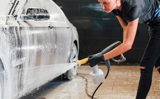 utilizando agua a alta presión. el automóvil negro moderno es limpiado por una mujer dentro de la estación de lavado de autos foto