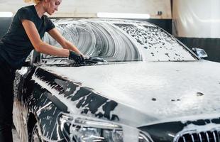 Wipes vehicle that is in white soap. Modern black automobile get cleaned by woman inside of car wash station photo