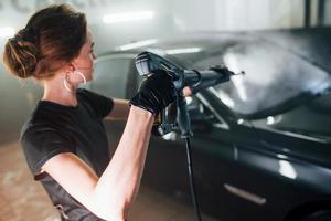 Using equipment with high pressure water. Modern black automobile get cleaned by woman inside of car wash station photo