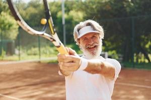 Playing game. Senior modern stylish man with racket outdoors on tennis court at daytime photo