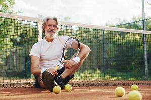 Sits on the ground and takes break. Senior modern stylish man with racket outdoors on tennis court at daytime photo