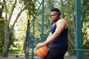 African american man with wireless headphones takes a break and leaning on the metal mesh with ball on the court outdoors photo
