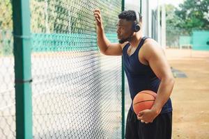 African american man with wireless headphones takes a break and leaning on the metal mesh with ball on the court outdoors photo