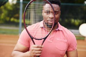 hombre afroamericano en camisa rosa de pie con racjet de tenis en la cancha al aire libre foto