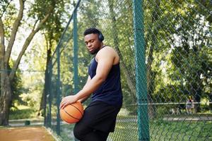 African american man with wireless headphones takes a break and leaning on the metal mesh with ball on the court outdoors photo