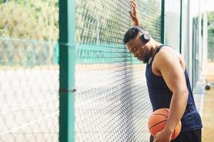 African american man with wireless headphones takes a break and leaning on the metal mesh with ball on the court outdoors photo