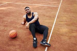 Tired african american man sits on the ground with ball on the court outdoors. Takes a break photo