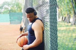 hombre afroamericano con auriculares inalámbricos toma un descanso y se apoya en la malla metálica con pelota en la cancha al aire libre foto