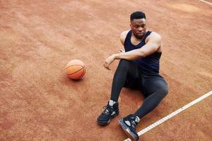 Tired african american man sits on the ground with ball on the court outdoors. Takes a break photo