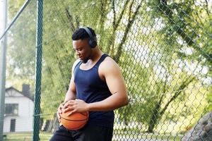 African american man with wireless headphones takes a break and leaning on the metal mesh with ball on the court outdoors photo