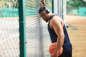 African american man with wireless headphones takes a break and leaning on the metal mesh with ball on the court outdoors photo