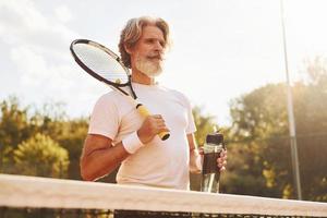 sosteniendo la raqueta. Senior hombre moderno y elegante con raqueta al aire libre en la cancha de tenis durante el día foto
