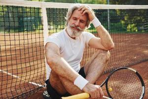 Sits on the ground and takes break. Senior modern stylish man with racket outdoors on tennis court at daytime photo