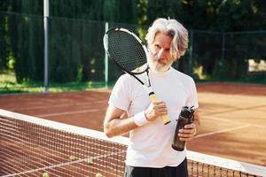 sosteniendo una botella de agua. Senior hombre moderno y elegante con raqueta al aire libre en la cancha de tenis durante el día