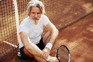 Sits on the ground and takes break. Senior modern stylish man with racket outdoors on tennis court at daytime photo