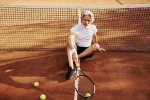 Sits on the ground and takes break. Senior modern stylish man with racket outdoors on tennis court at daytime photo