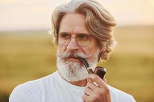 retrato de un anciano elegante con cabello gris y barba que está parado al aire libre en el campo en un día soleado y fumando foto