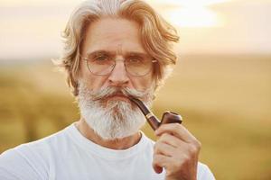 retrato de un anciano elegante con cabello gris y barba que está parado al aire libre en el campo en un día soleado y fumando foto