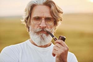 retrato de un anciano elegante con cabello gris y barba que está parado al aire libre en el campo en un día soleado y fumando foto